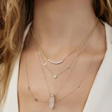 A close-up of a woman’s neck wearing four yellow gold necklaces - two Custom Gold Necklaces (one with three charms and the other with four charms), the Clarity Retreat Necklace and the Diamond Arc Layering Necklace, with a cream top.