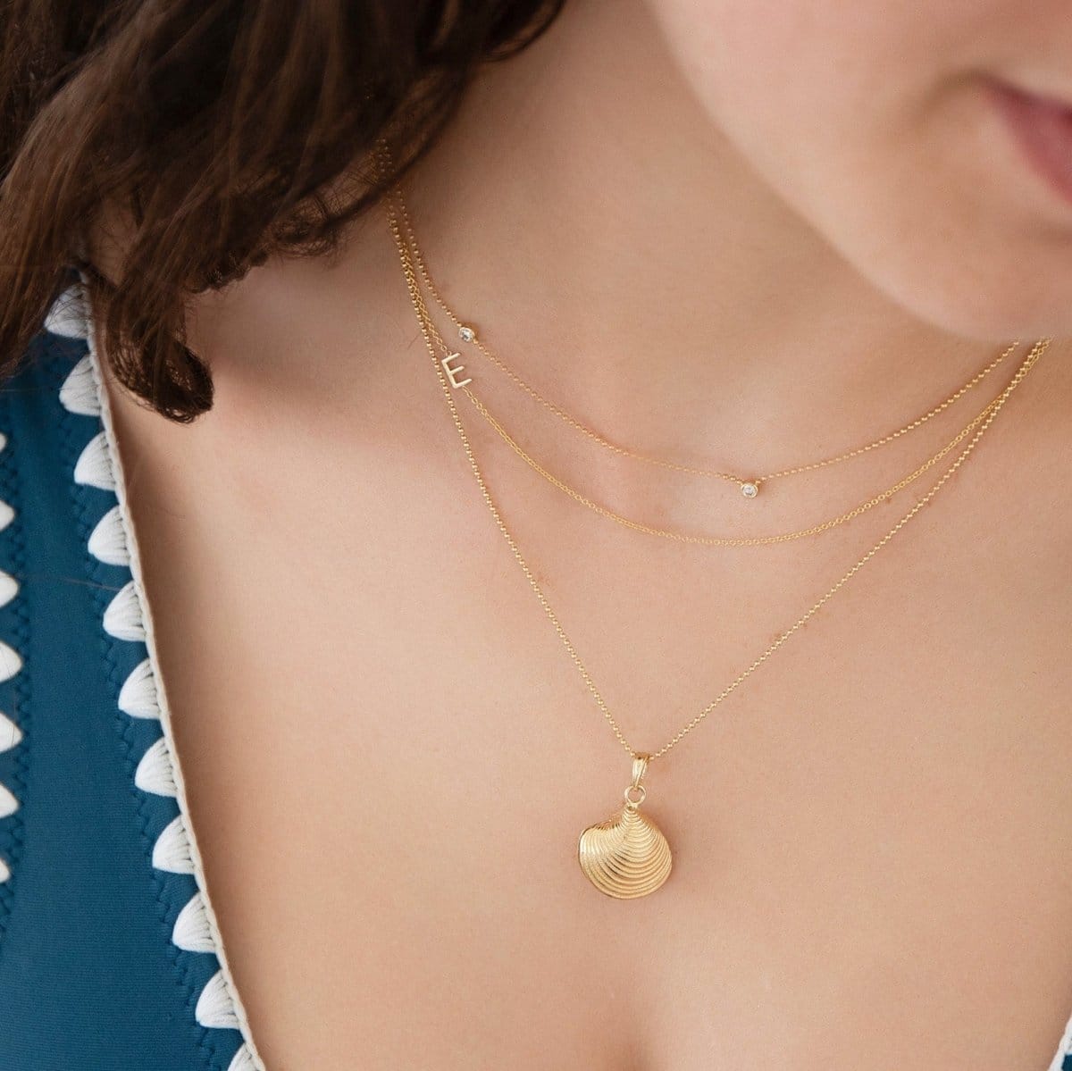Close-up of a woman wearing a Triple Diamond Layering Necklace, 14k Gold Asymmetrical Letter Necklace - E and Classic Bead Chain with a Golden Coast Shell. 