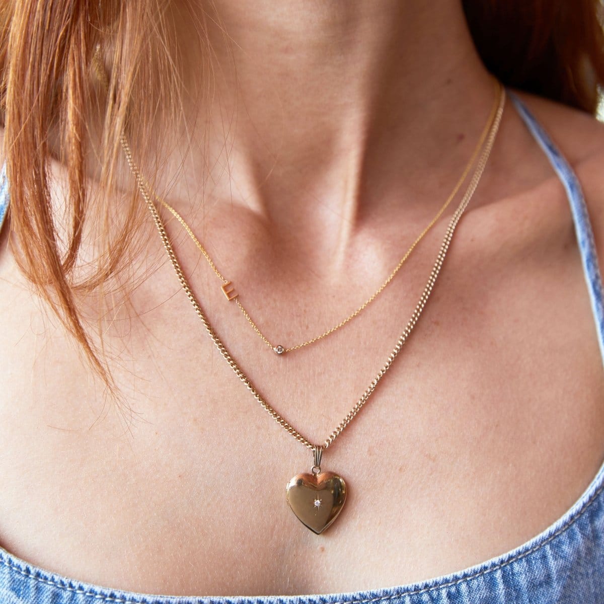 A close-up of a woman’s neck wearing a Monogram Necklace with Diamond and the letter E and a Round Curb Chain with a Diamond Heart Locket Charm, all in yellow gold, wearing a denim top. 