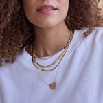 Close-up of a woman's neck wearing The Ultimate Diamond Necklace, a Bold Link Chain, a Sequence Chain and a Heart Necklace, all in yellow gold, with a white-shirt and her brown curly hair visible. 