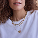 Close-up of a woman's neck wearing The Ultimate Diamond Necklace, a Bold Link Chain, a Sequence Chain and a Heart Necklace, all in yellow gold, with a white-shirt and her brown curly hair visible. 