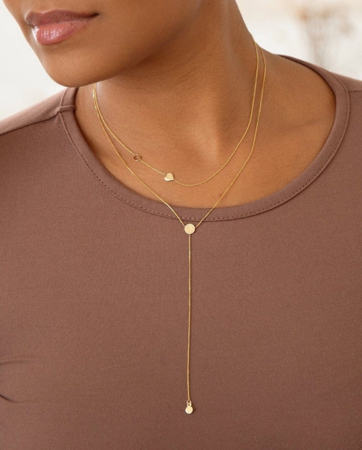 A close-up of a woman wearing a brown top with two necklaces, being a Monogram Necklace with Heart Yellow Gold and the letter C charm, and a Gold Lariat Necklace. 
