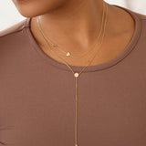 A close-up of a woman wearing a brown top with two necklaces, being a Monogram Necklace with Heart Yellow Gold and the letter C charm, and a Gold Lariat Necklace. 