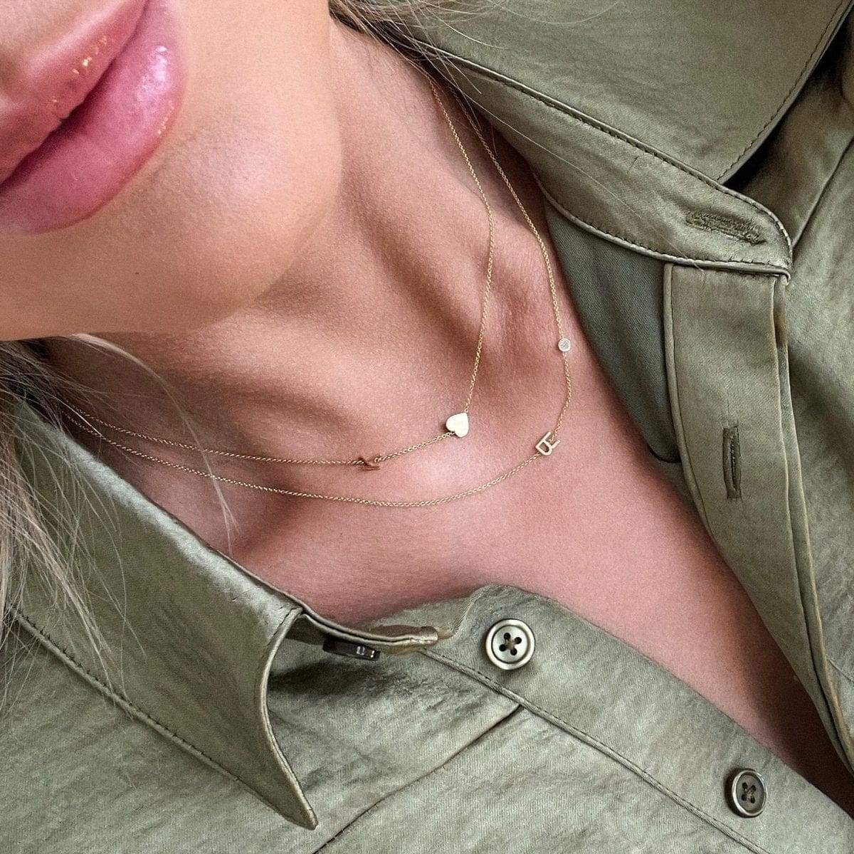 A close-up of a woman in a green shirt wearing a Monogram Necklace with Heart Yellow Gold and a letter K charm, and a Monogram Necklace with Diamond Yellow Gold and a letter R charm.