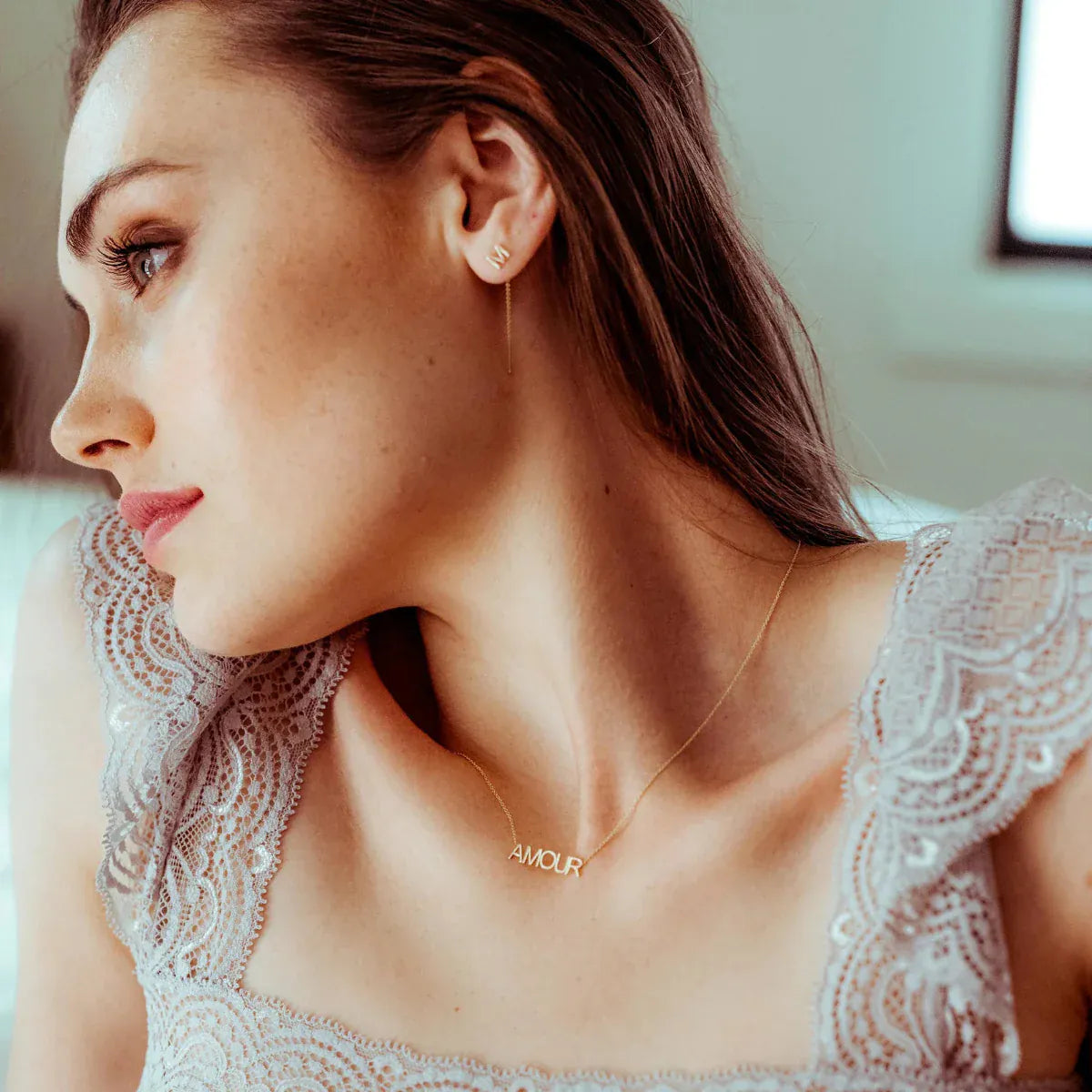 A woman wearing a 14K Gold Letter Threader Earring 'M' and an AMOUR Necklace, both in yellow gold, with a light grey lace top and a blurred background.