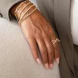 A woman's hand wearing a yellow gold Criss-Cross Diamond Daze Ring, Rolo Bracelet, Element Short Link Chain, Bold Link Bracelet, Down the Line Bracelet and Love Set Match Bracelet against a cream fabric surface, styled in a white top and khaki jacket. 
