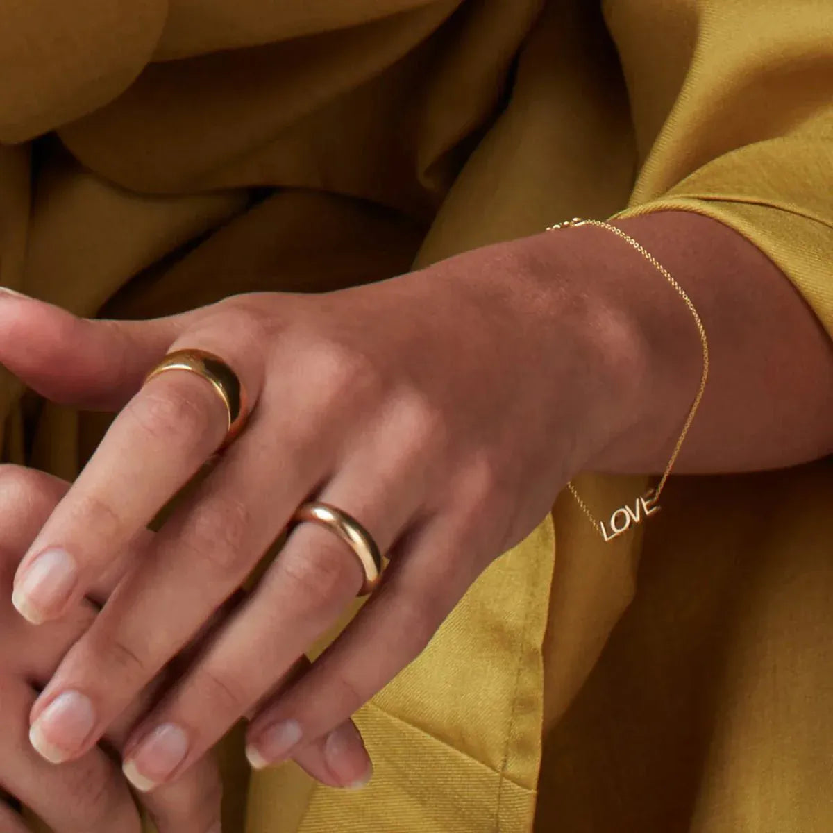 A close-up of a woman's hand and arm wearing a Love Bracelet, a Thick Band and a Tube Ring, in mustard clothing. 
