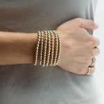 A close up a person's wrist wearing multiple stacked Luxe Bubble Bracelets and gold rings, with a sage green top. 