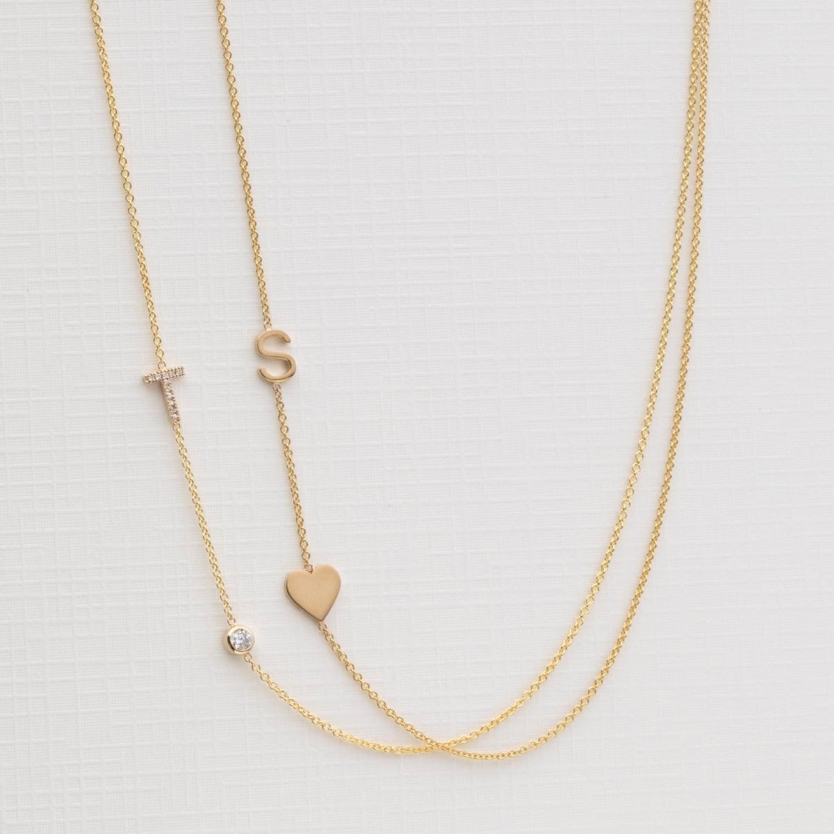 A Monogram Necklace with Heart Yellow Gold and a letter S charm, and a Pavé Monogram Necklace with Diamond and a letter T charm against an off-white background. 