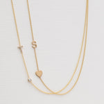 A Monogram Necklace with Heart Yellow Gold and a letter S charm, and a Pavé Monogram Necklace with Diamond and a letter T charm against an off-white background. 