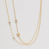 A Monogram Necklace with Heart Yellow Gold and a letter S charm, and a Pavé Monogram Necklace with Diamond and a letter T charm against an off-white background. 