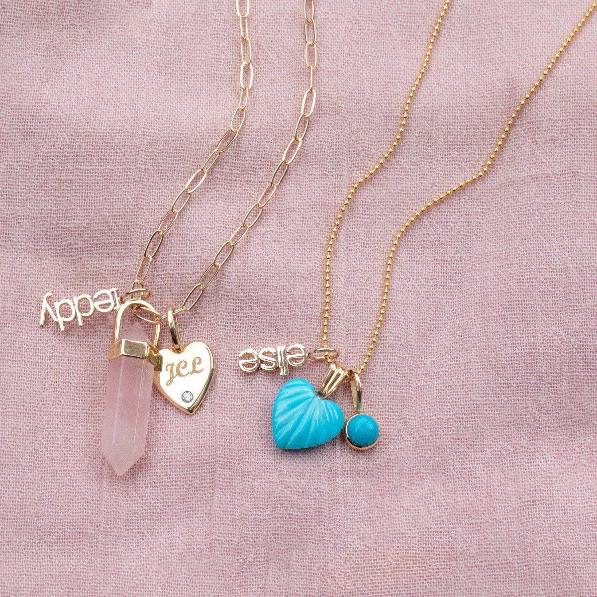 A yellow gold Element Short Link Chain with a Petite Nameplate Pendant in 'teddy', a Rose Quartz Balance Crystal Charm and an Engraved Heart Charm, and a yellow gold Classic Bead Chain with a Petite Nameplate Pendant in 'elise', an Etched Turquoise Heart Charm and a Turquoise Dangle Charm, against a light pink fabric background.