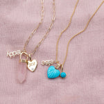 A yellow gold Element Short Link Chain with a Petite Nameplate Pendant in 'teddy', a Rose Quartz Balance Crystal Charm and an Engraved Heart Charm, and a yellow gold Classic Bead Chain with a Petite Nameplate Pendant in 'elise', an Etched Turquoise Heart Charm and a Turquoise Dangle Charm, against a light pink fabric background.