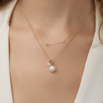 A close-up of a woman’s neck wearing a Pavé Monogram Necklace with Diamond and an H charm in yellow gold, a Classic Bead Chain with a Perla Pendant and a Duchess Diamond Pendant, in a cream top.