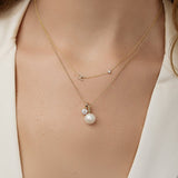 A close-up of a woman’s neck wearing a Pavé Monogram Necklace with Diamond and an H charm in yellow gold, a Classic Bead Chain with a Perla Pendant and a Duchess Diamond Pendant, in a cream top.