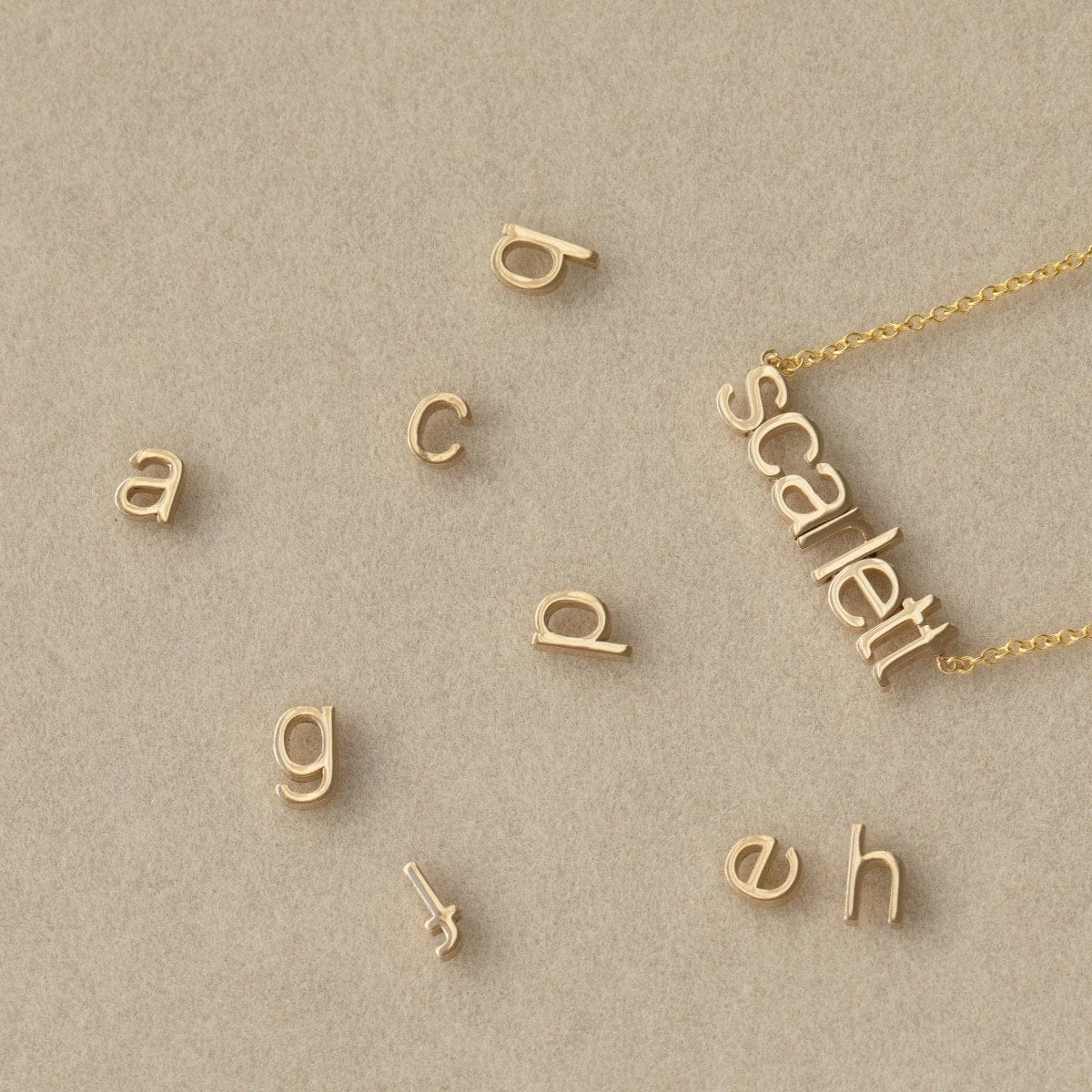 A Petite Nameplate Necklace in Scarlett with several Petite Letter Charms, all in yellow gold, against a light beige background. 
