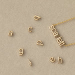A Petite Nameplate Necklace in Scarlett with several Petite Letter Charms, all in yellow gold, against a light beige background. 