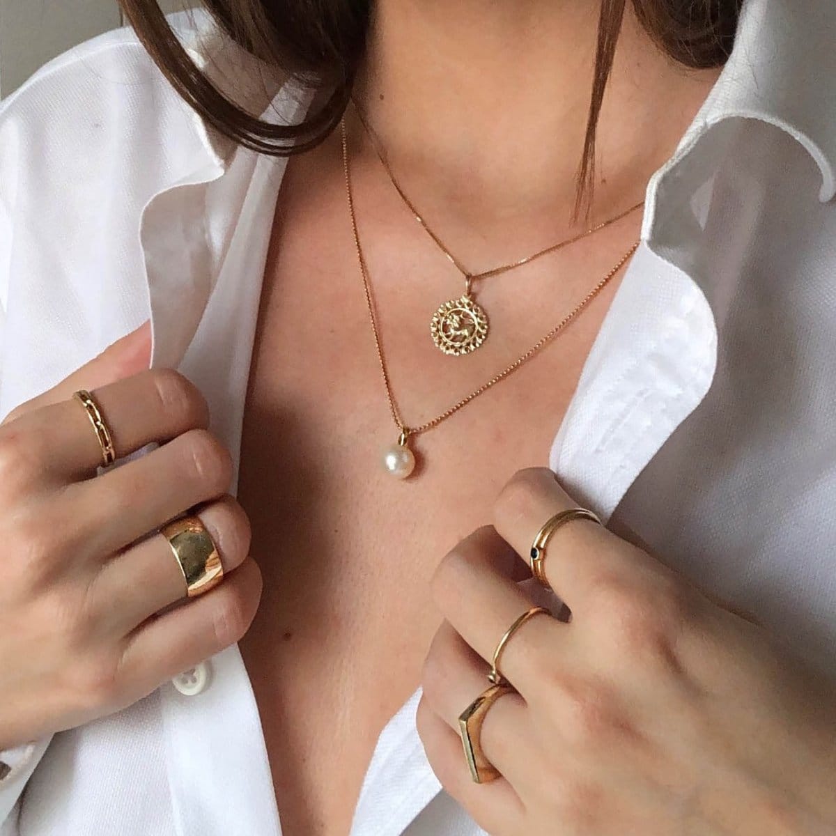 Close-up of a brunette woman's décolletage wearing a Zodiac Necklace - Leo and a Perla Necklace, with her hands wearing a Link Ring, a Thick Band, Birthstone Rings, and a Geo Stacking Ring Square, in a white shirt.