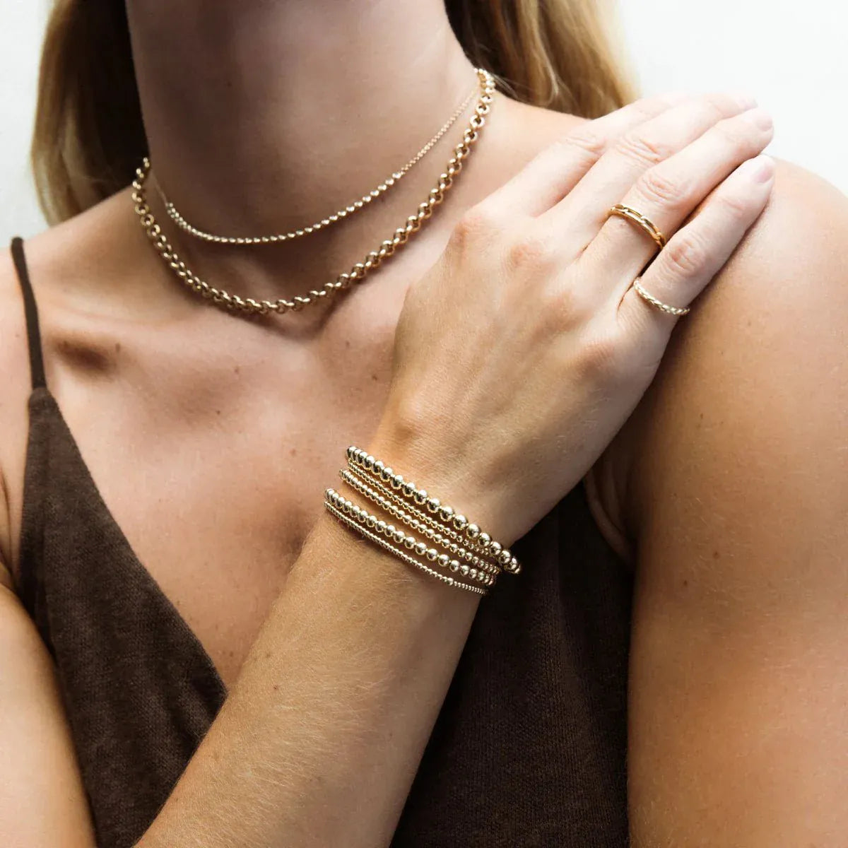 A close-up of a woman’s décolletage and arm wearing a Luxe Bubble Bracelet Set, a Link Ring, a Braided Ring, a Rolo Necklace and The Everyday Diamond Necklace, all in yellow gold, with a brown top.