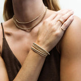 A close-up of a woman’s décolletage and arm wearing a Luxe Bubble Bracelet Set, a Link Ring, a Braided Ring, a Rolo Necklace and The Everyday Diamond Necklace, all in yellow gold, with a brown top.