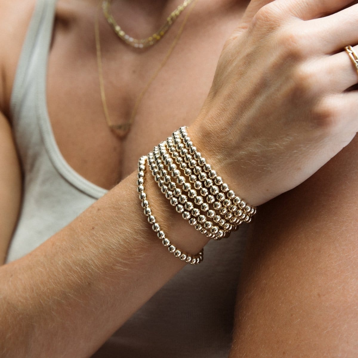 Close-up of a woman’s arm wearing multiple Statement Bubble Bracelets, gold necklaces and a Link Ring, in a light sage green top.