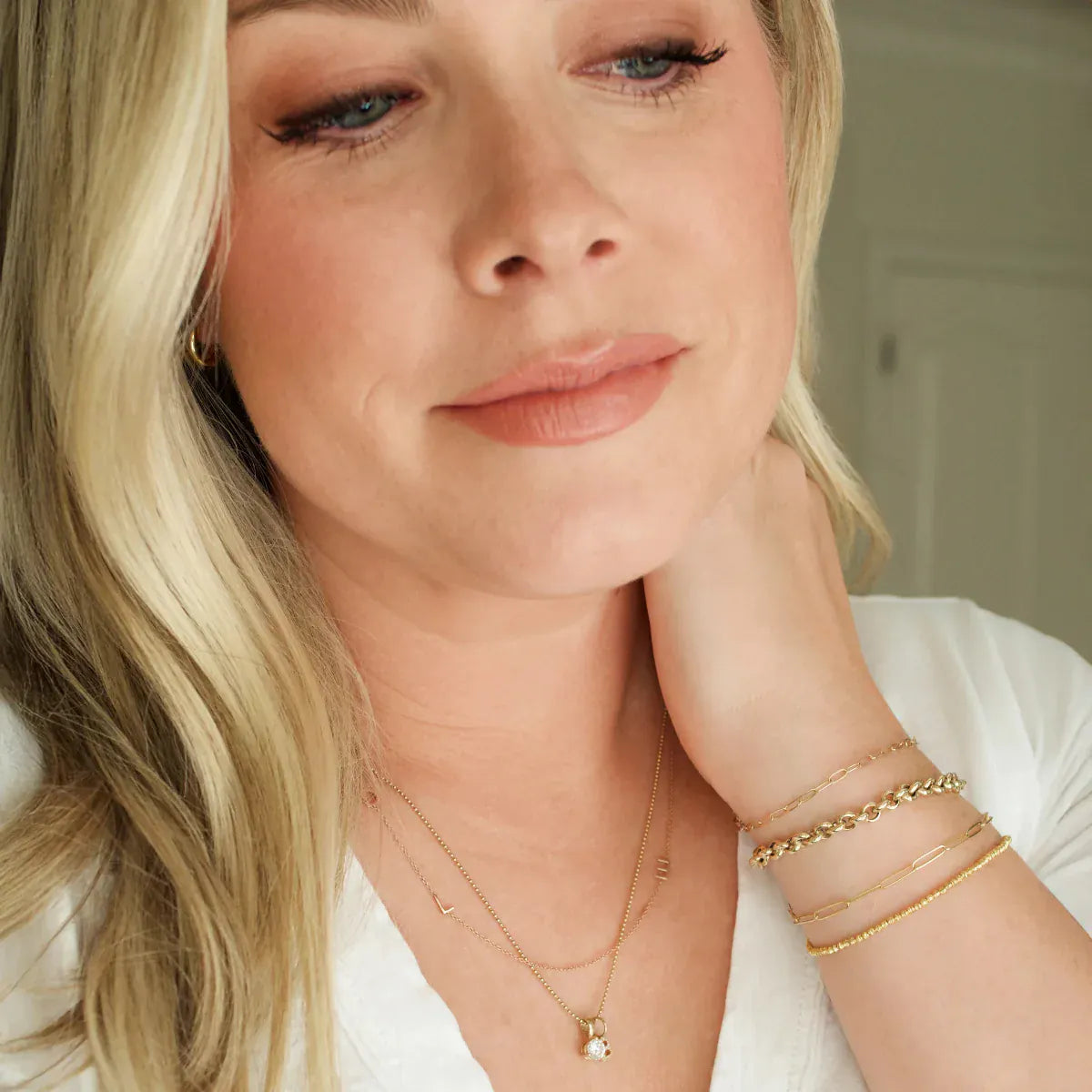 A woman wearing a Custom Gold Necklace - 3 Letters, a Classic Bead Chain with a Princess Diamond Pendant, an Element Short Link Chain, Element Long Link Chain, Rolo Bracelet and Diamond Cut Bracelet, all in yellow gold, with blonde hair and a white top against a blurred background. 
