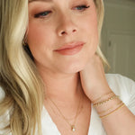 A woman wearing a Custom Gold Necklace - 3 Letters, a Classic Bead Chain with a Princess Diamond Pendant, an Element Short Link Chain, Element Long Link Chain, Rolo Bracelet and Diamond Cut Bracelet, all in yellow gold, with blonde hair and a white top against a blurred background. 