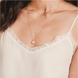 A close-up of a woman’s chest wearing a cream lace top and three yellow gold necklaces: a Perla Necklace, an Angel Wing Charm Necklace and a Custom Gold Necklace with four charms (three birthstones and a Star of David).