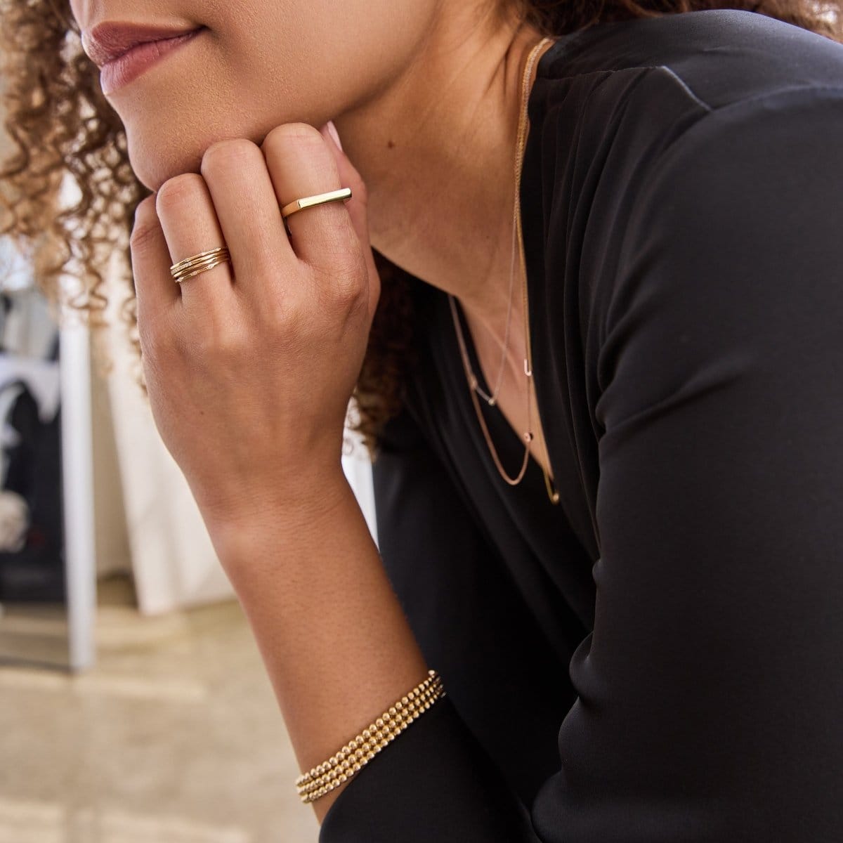 Close-up of a woman wearing a yellow gold Geo Stacking Ring | Square, Delicate Band, Pave Diamond Ring, two 4 Diamond Rings and three Bubble Bracelets with a black top, against a softly blurred background.  