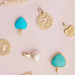 Several yellow gold and turquoise pendants and charms, including Zodiac Pendants, Petite Nameplate Pendants, a Perla Pendant, a Turquoise Heart Charm, an Etched Turquoise Heart Charm and an Engraved Heart Charm on a light pink background. 