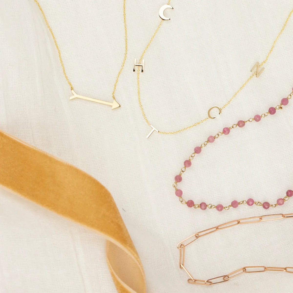 A Straight as an Arrow Necklace in yellow gold, a Custom Gold Necklace - 5 Letters  in yellow gold, The Peony Bouquet Necklace and an Element Long Link Chain in rose gold on a white fabric background with a yellow piece of ribbon. 