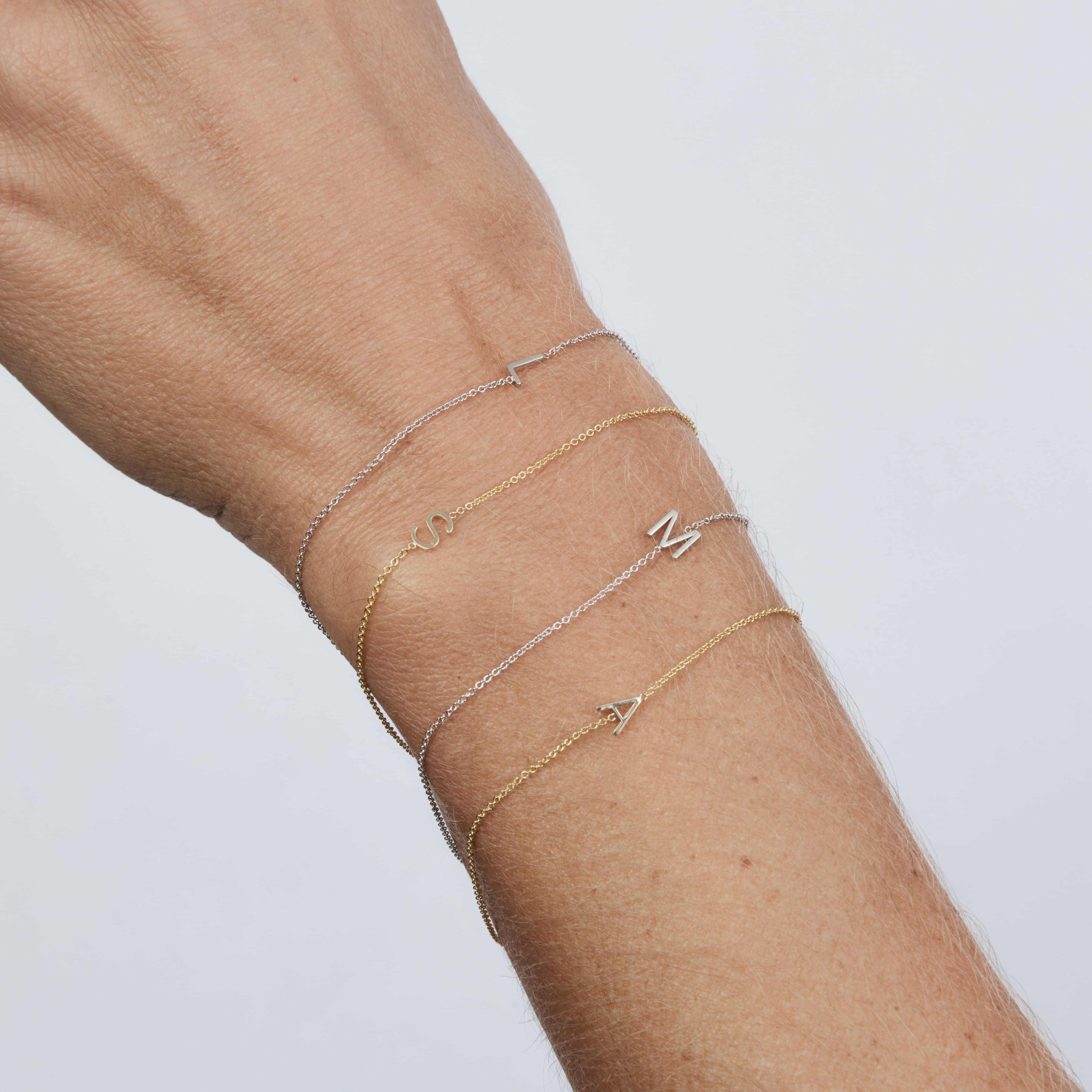 Gold bracelets with letter charms on a wrist against a light gray background