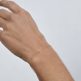 A delicate gold chain bracelet with a small open D-shaped charm, shown worn on a person's wrist against a light gray background.
