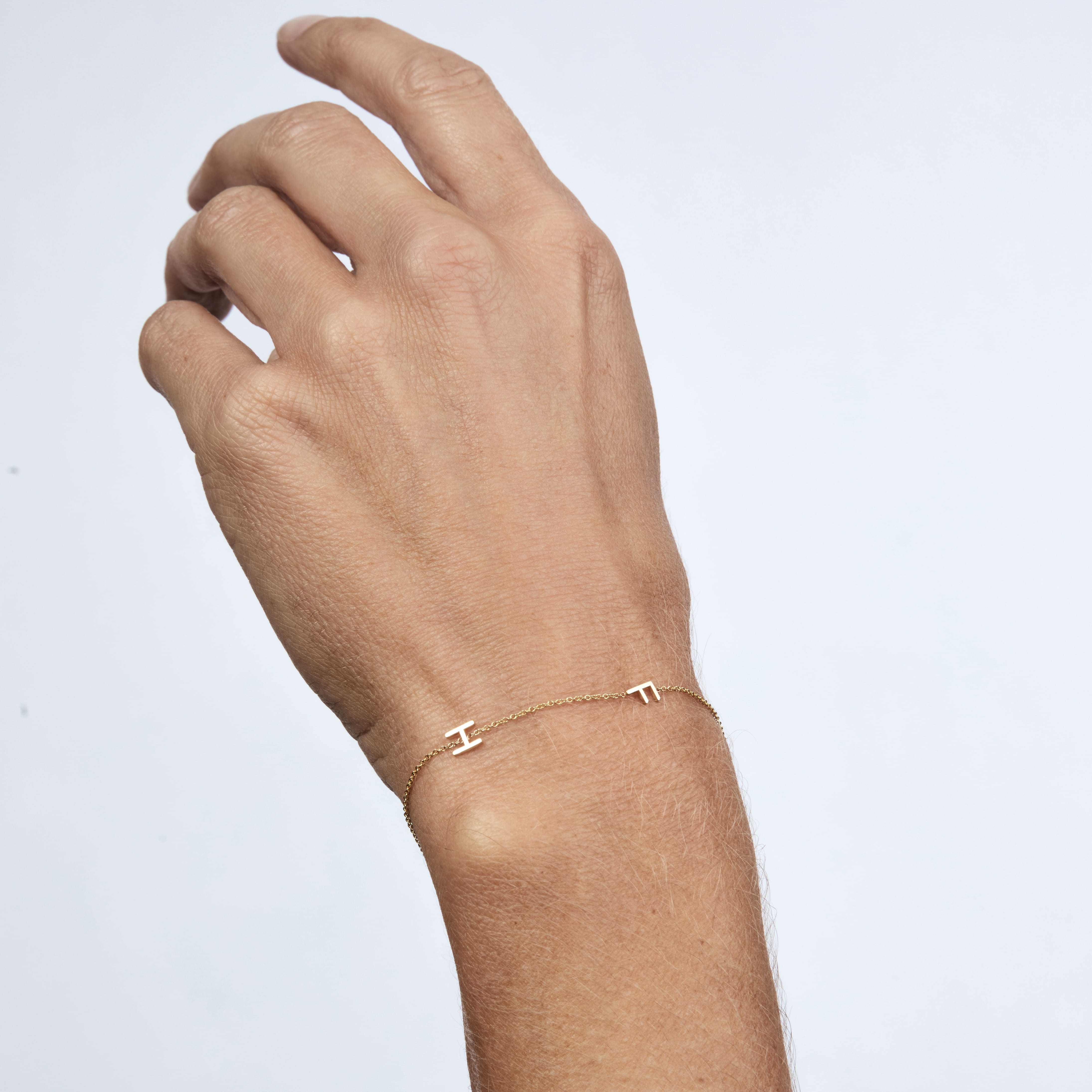 A delicate rose gold chain bracelet with a small Greek letter 'H' charm, shown worn on a person's wrist against a clean, light gray background.