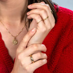 Close-up of a brunette woman wearing a Zodiac Necklace - Cancer, a Link Ring, a Tube Ring,and a Delicate Band, all in yellow gold, with a red knit against a light grey background.