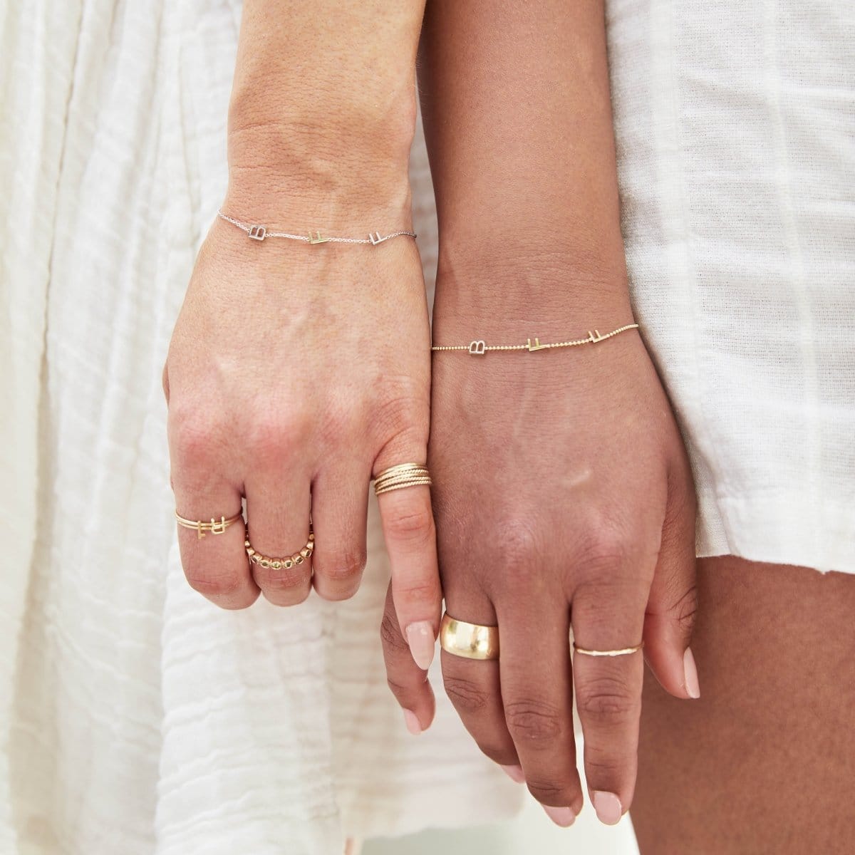 Close-up of two hands, each wearing a BFF Bracelet and several yellow gold rings, including a 14K Gold Letter Ring in A, a Lucky Number Ring in 1, a Hydrangea Bouquet Ring, a Thick Band, Delicate Bands, Delicate Twist Bands and a 4 Diamond Ring. 