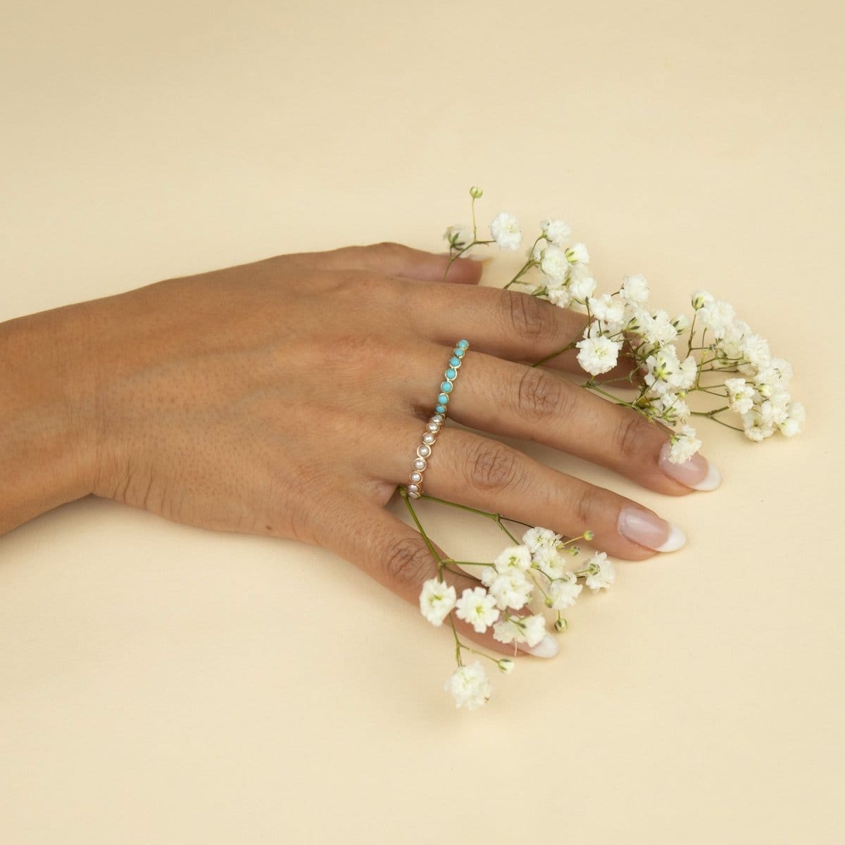A hand wearing The Bluebell Bouquet Ring and The Lily Bouquet Ring holding small white flowers against a light yellow background. 