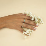 A hand wearing The Bluebell Bouquet Ring and The Lily Bouquet Ring holding small white flowers against a light yellow background. 
