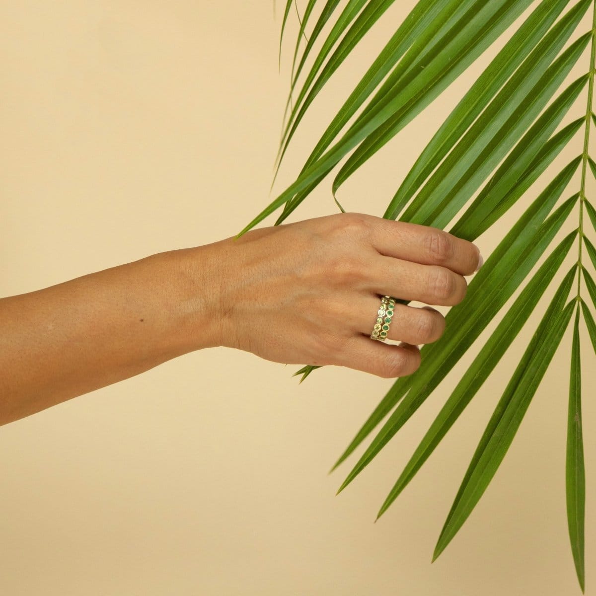 A hand wearing The Fern Bouquet Ring and The Evermore Ring holding a green plant against a light yellow background. 