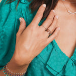 Close-up of a woman's hand wearing rose gold Delicate Trio Stacking Rings, an Open Diamond Ring, several Bouquet Bracelets, Diamond Arc Layering Necklace, styled in a green blue blouse. 