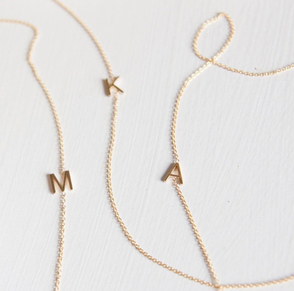Three gold necklaces with single letter charms, being M, K and A on a plain background. 