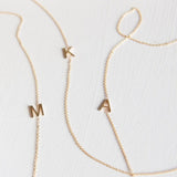 Three gold necklaces with single letter charms, being M, K and A on a plain background. 