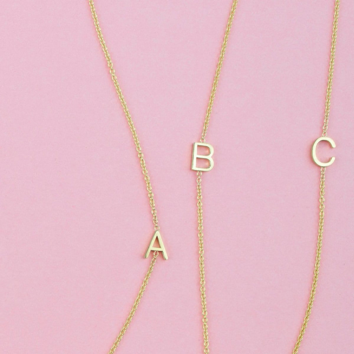 Three gold necklaces with single letter charms A, B and C on a pink background. 