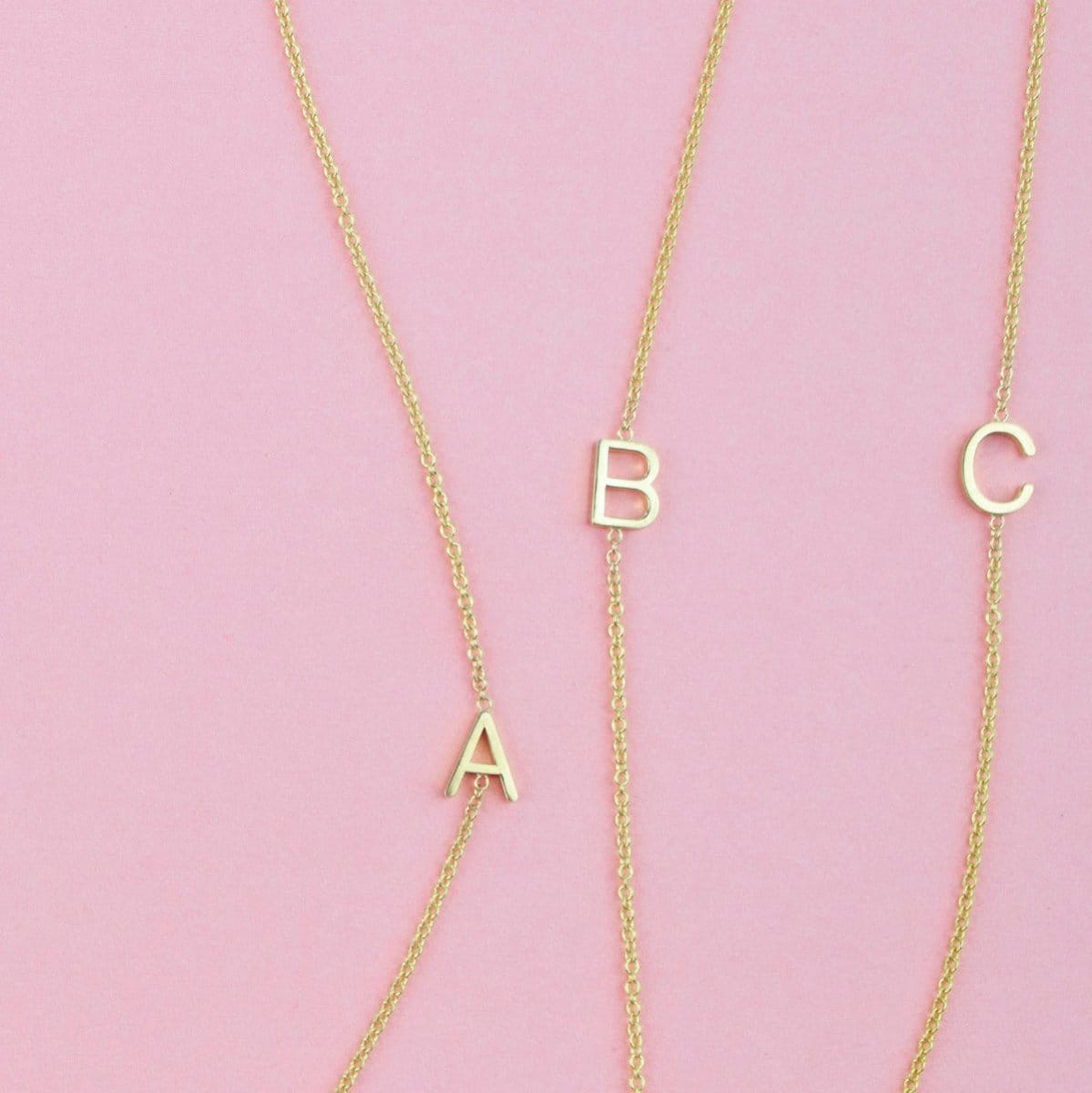 Three gold necklaces with single letter charms A, B and C on a pink background. 