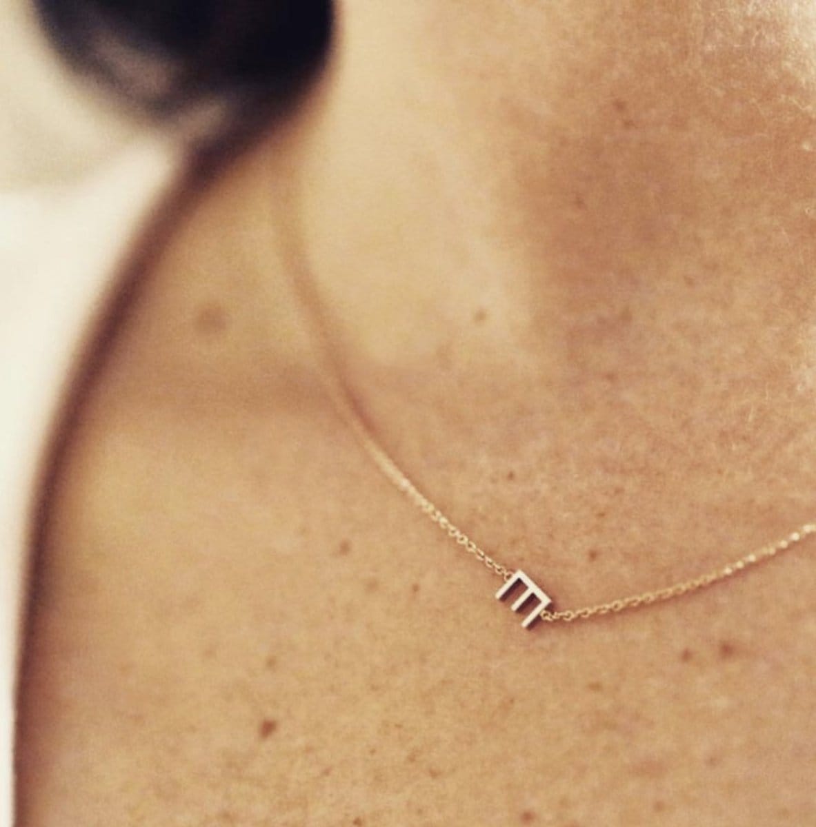 A close-up of a woman's neck wearing a gold necklace with a letter E charm. 