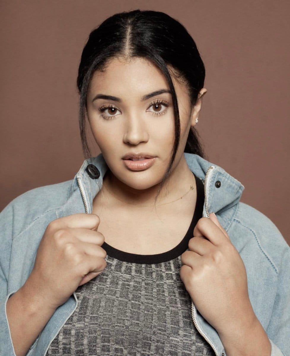 A woman wearing a dark grey top and denim jacket, a necklace with a letter W charm and stud earrings.