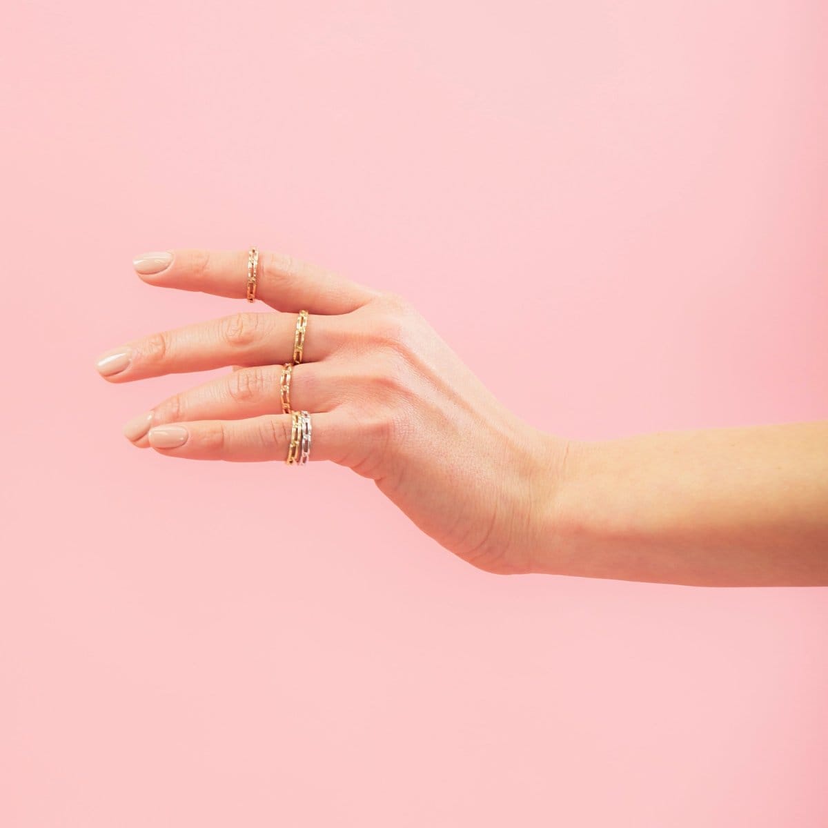 A hand wearing several Link Rings in yellow gold, rose gold and white gold against a light pink background. 