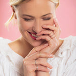 A woman wearing several rings, including mixed metal Flat Rings, with blonde hair and a white top, against a light pink background. 