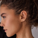 A close-up of a woman's side profile wearing two earrings - a mini letter E gold stud and a diamond stud. 