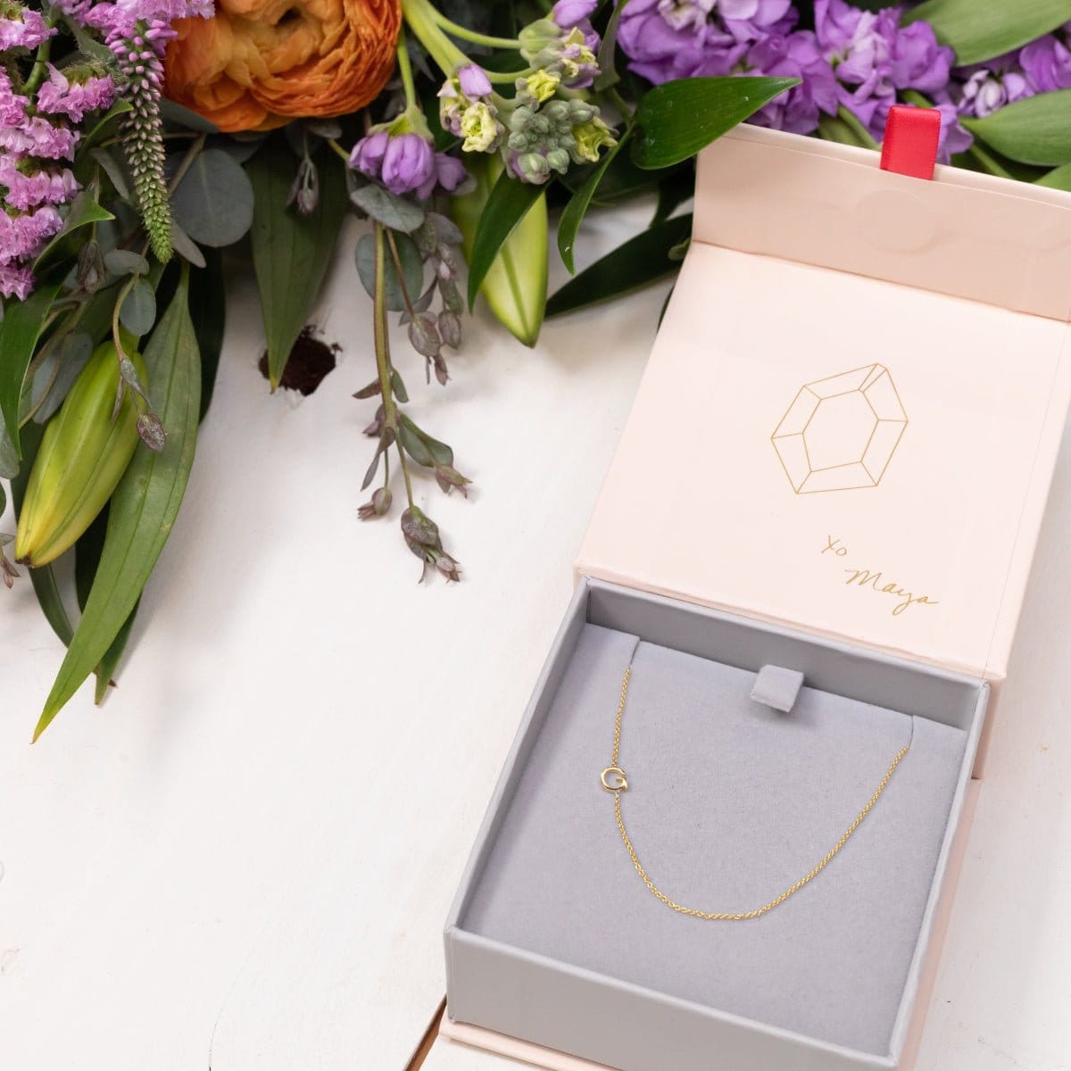 A gold necklace with a letter G charm in a jewellery box, with flowers in the background.